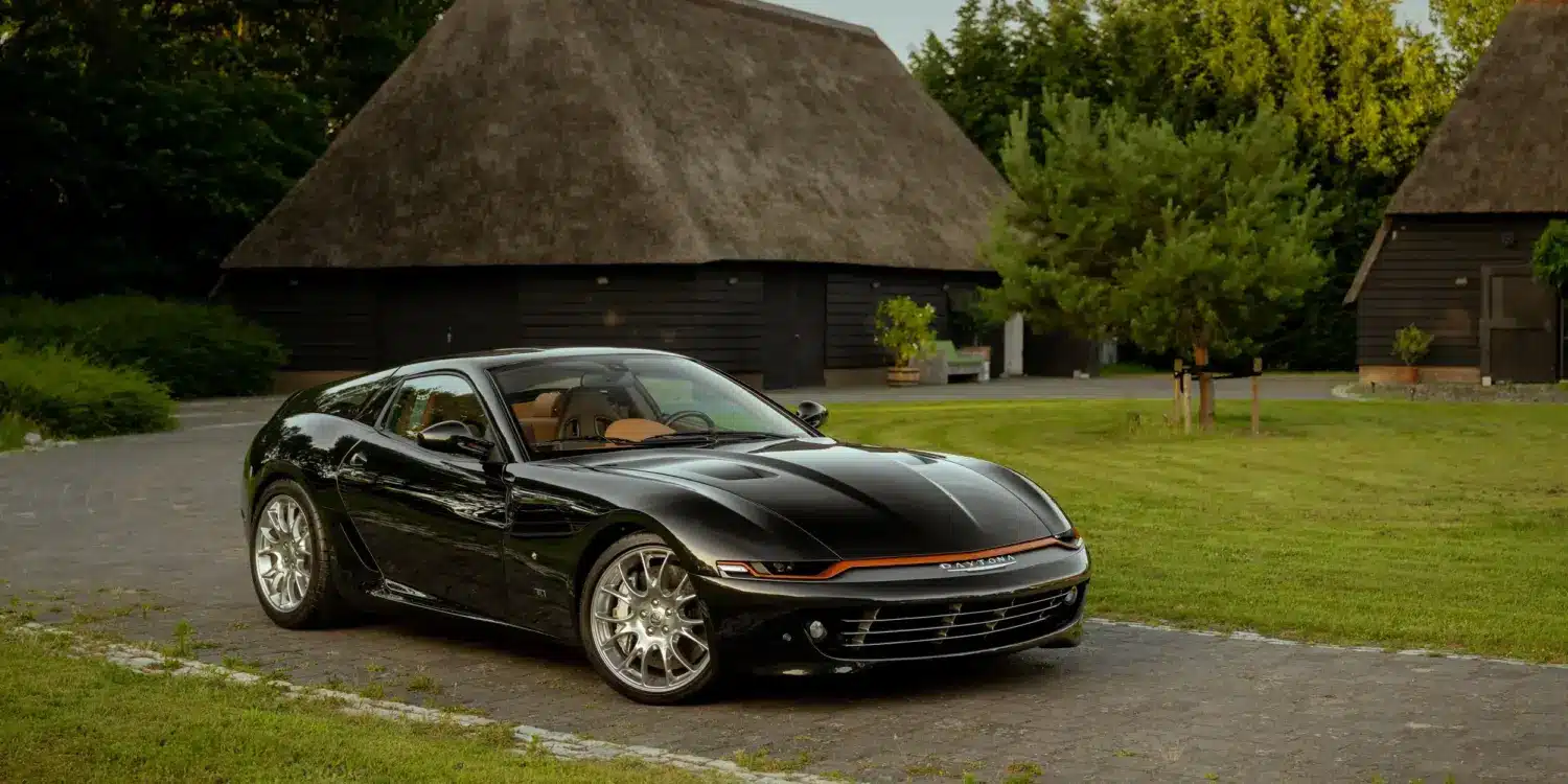 Schwarzer Daytona Shooting Brake Hommage parkt auf einer Auffahrt vor traditionellen strohgedeckten Gebäuden.