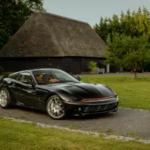 Schwarzer Daytona Shooting Brake Hommage parkt auf einer Auffahrt vor traditionellen strohgedeckten Gebäuden.