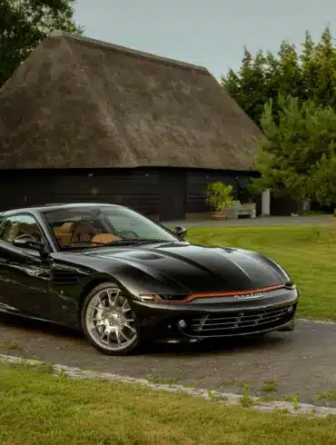 Schwarzer Daytona Shooting Brake Hommage parkt auf einer Auffahrt vor traditionellen strohgedeckten Gebäuden.