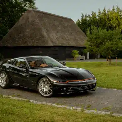 Schwarzer Daytona Shooting Brake Hommage parkt auf einer Auffahrt vor traditionellen strohgedeckten Gebäuden.