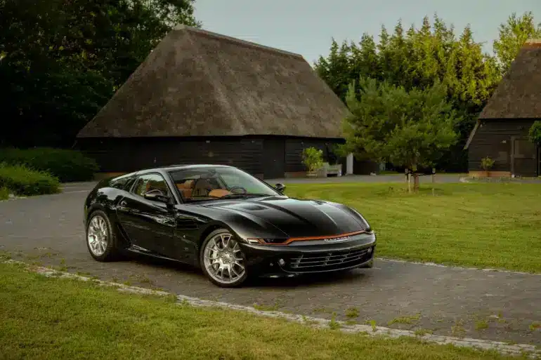 Schwarzer Daytona Shooting Brake Hommage parkt auf einer Auffahrt vor traditionellen strohgedeckten Gebäuden.