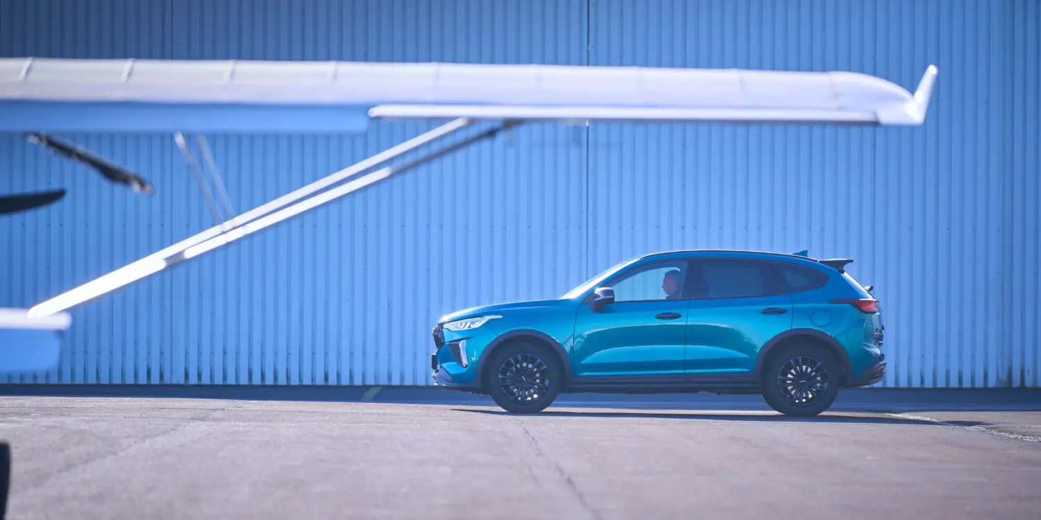 Seitliche Ansicht des GWM HAVAL Jolion Pro in Blau, geparkt auf einem Flugplatz mit Flugzeug im Vordergrund.