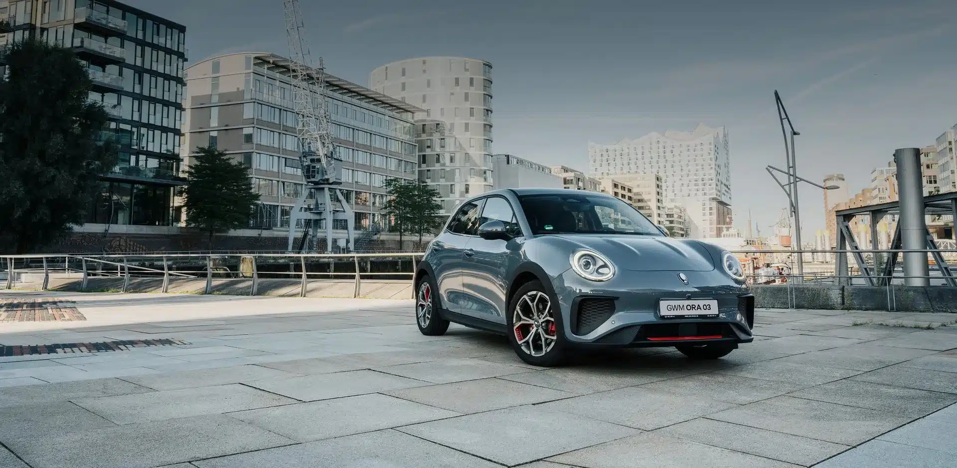 GWM ORA 03 Elektroauto in Grau, abgestellt auf einem urbanen Platz mit modernen Gebäuden im Hintergrund.