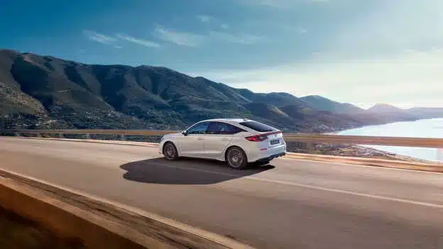 Weißer Honda Civic e:HEV fährt auf einer Straße mit Bergpanorama und Meer im Hintergrund.