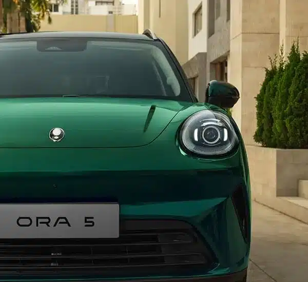 Front des GWM ORA 5 SUV mit grünem Lack und ovalen LED-Scheinwerfern vor urbaner Kulisse