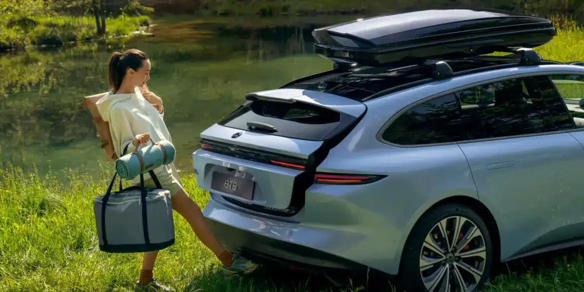 Eine Frau öffnet freihändig den Kofferraum eines silbernen NIO ET5 Touring, der mit Dachbox am Ufer eines Sees steht.