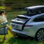 Eine Frau öffnet freihändig den Kofferraum eines silbernen NIO ET5 Touring, der mit Dachbox am Ufer eines Sees steht.
