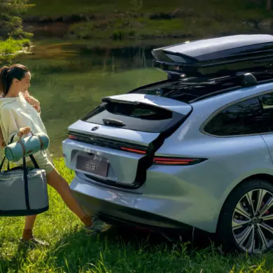 Eine Frau öffnet freihändig den Kofferraum eines silbernen NIO ET5 Touring, der mit Dachbox am Ufer eines Sees steht.