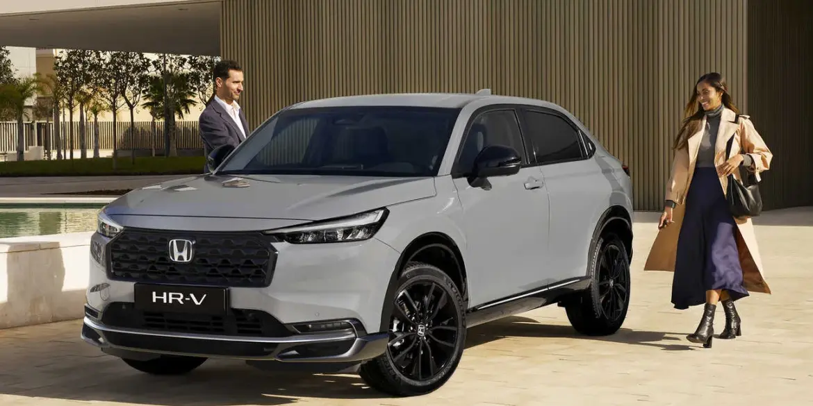 Honda HR-V e:HEV in Grau mit schwarzem Grill, geparkt vor moderner Architektur, zwei Personen daneben.