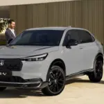 Honda HR-V e:HEV in Grau mit schwarzem Grill, geparkt vor moderner Architektur, zwei Personen daneben.