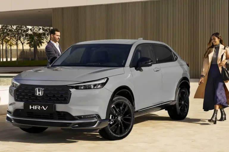 Honda HR-V e:HEV in Grau mit schwarzem Grill, geparkt vor moderner Architektur, zwei Personen daneben.