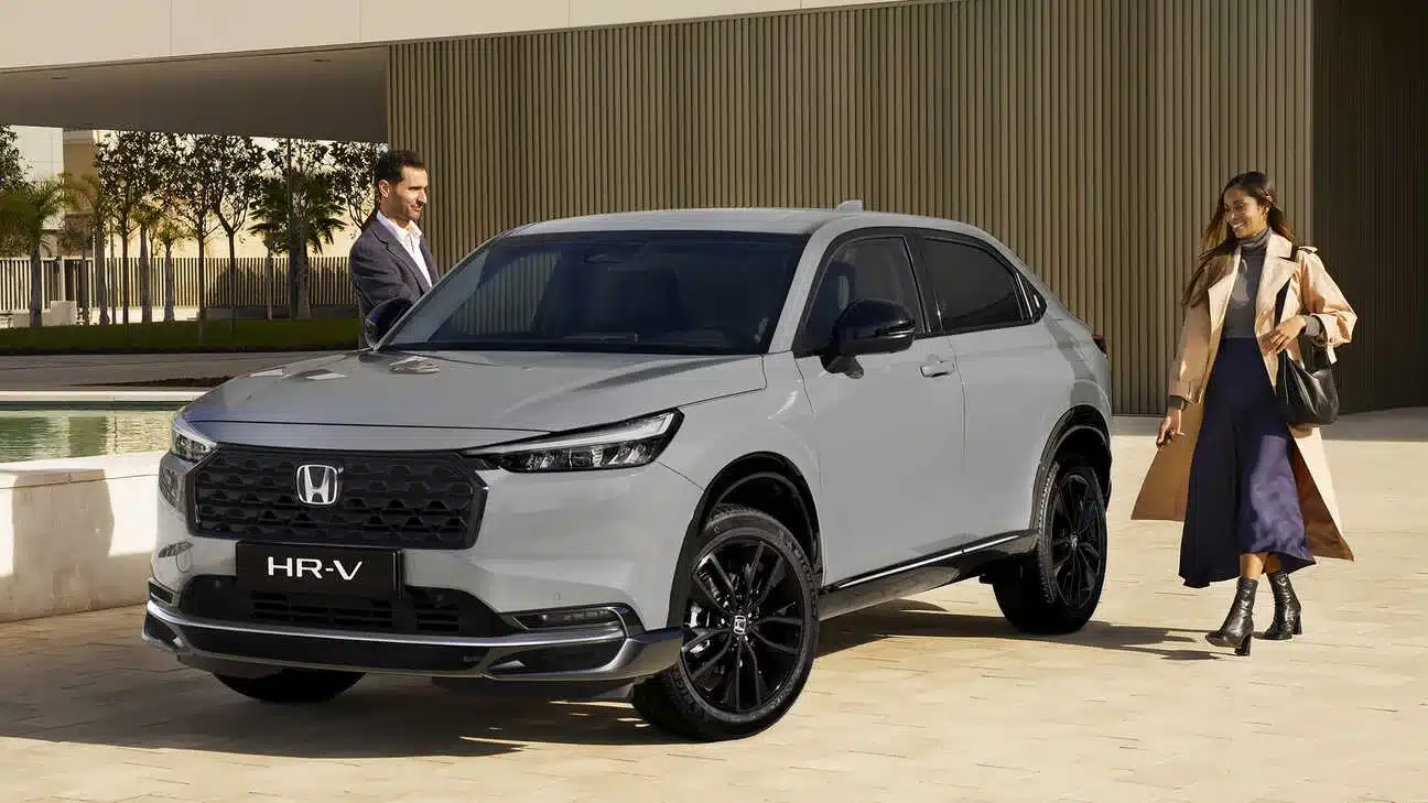 Honda HR-V e:HEV in Grau mit schwarzem Grill, geparkt vor moderner Architektur, zwei Personen daneben.