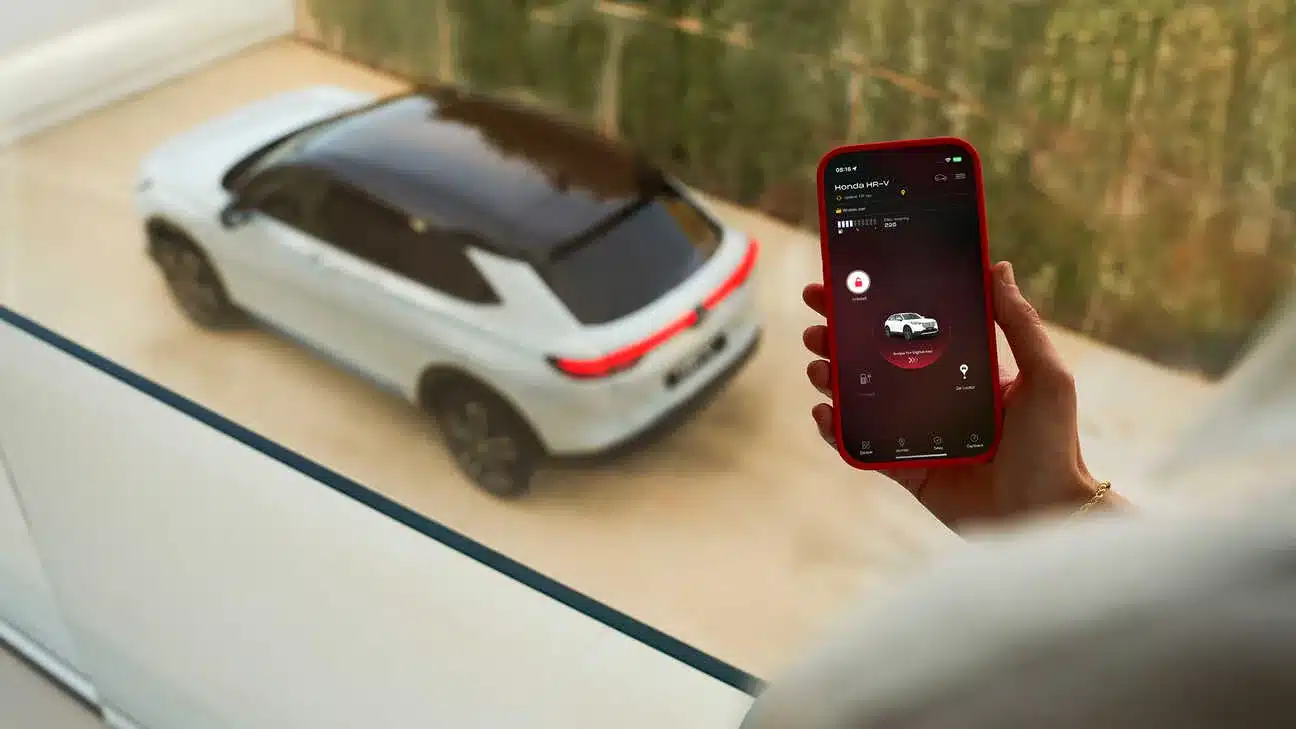 Frau hält Smartphone mit geöffneter My Honda+ App, im Hintergrund steht ein Honda HR-V e:HEV in einer Einfahrt.