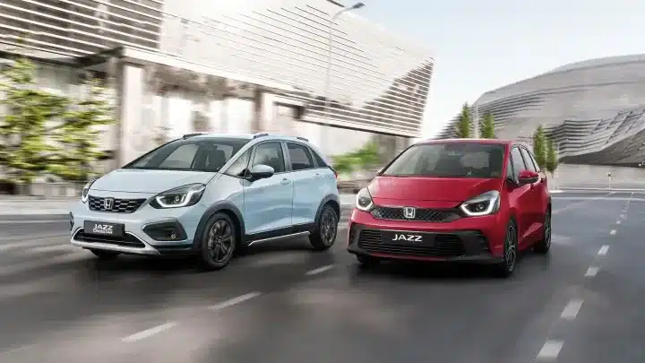 Zwei Honda Jazz e:HEV Modelle, einer in Hellblau (Crosstar Advance), einer in Rot, fahren nebeneinander auf einer städtischen Straße.