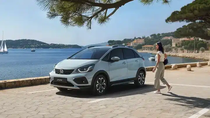 Honda Jazz e:HEV in heller Lackierung seitlich an einer Uferpromenade geparkt, eine Person läuft daran vorbei.