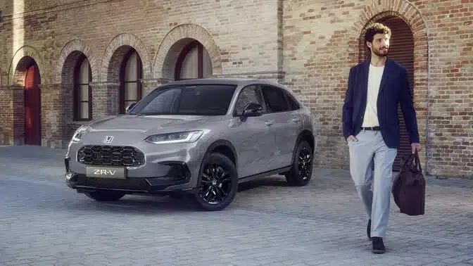 Honda ZR-V e:HEV Hybrid-SUV in Grau steht vor einer Backsteinmauer, ein Mann geht vorbei.