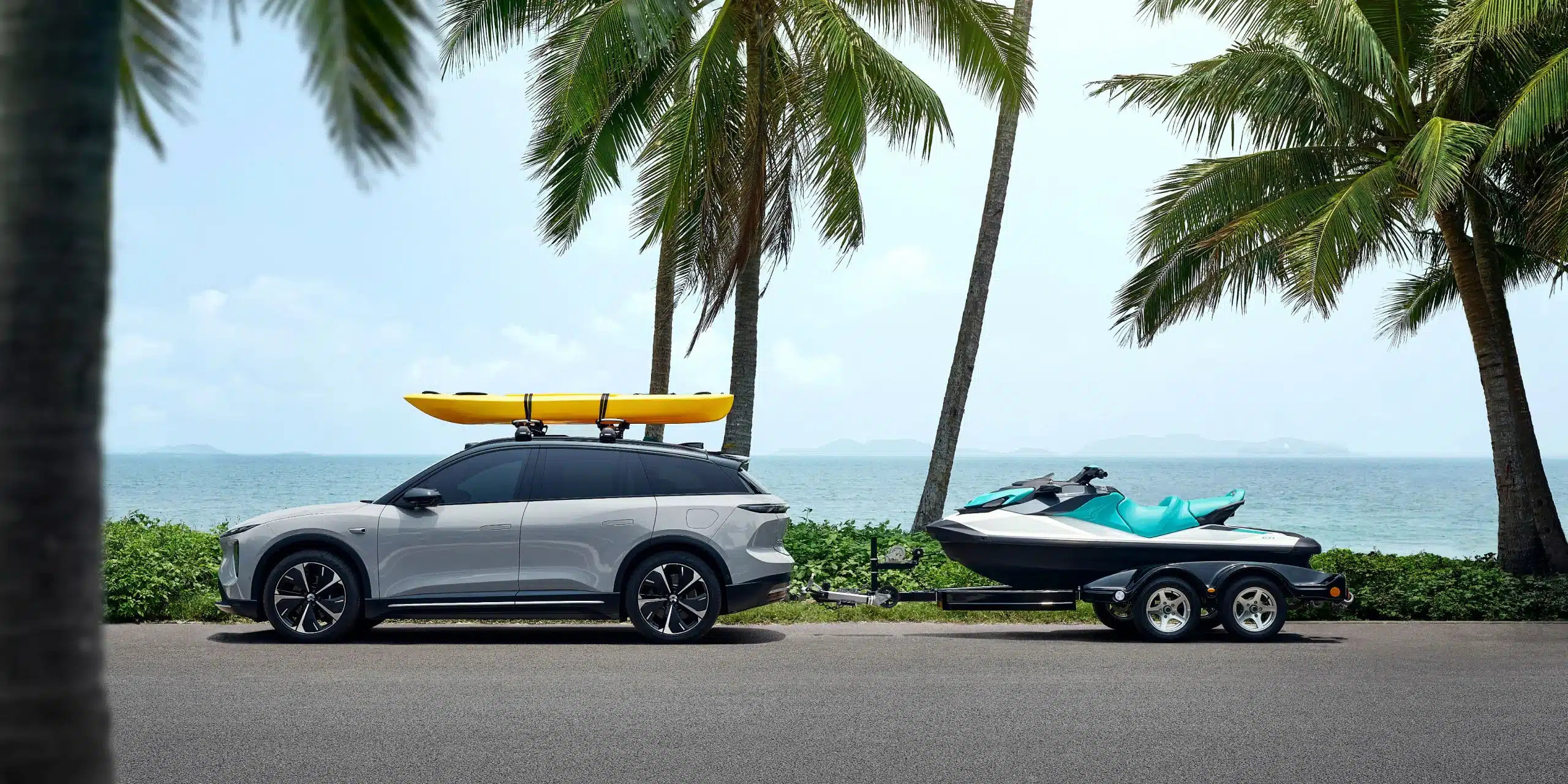 NIO EL6 Elektro-SUV mit Kajak auf dem Dach und Bootstrailer vor Palmen und Meer.