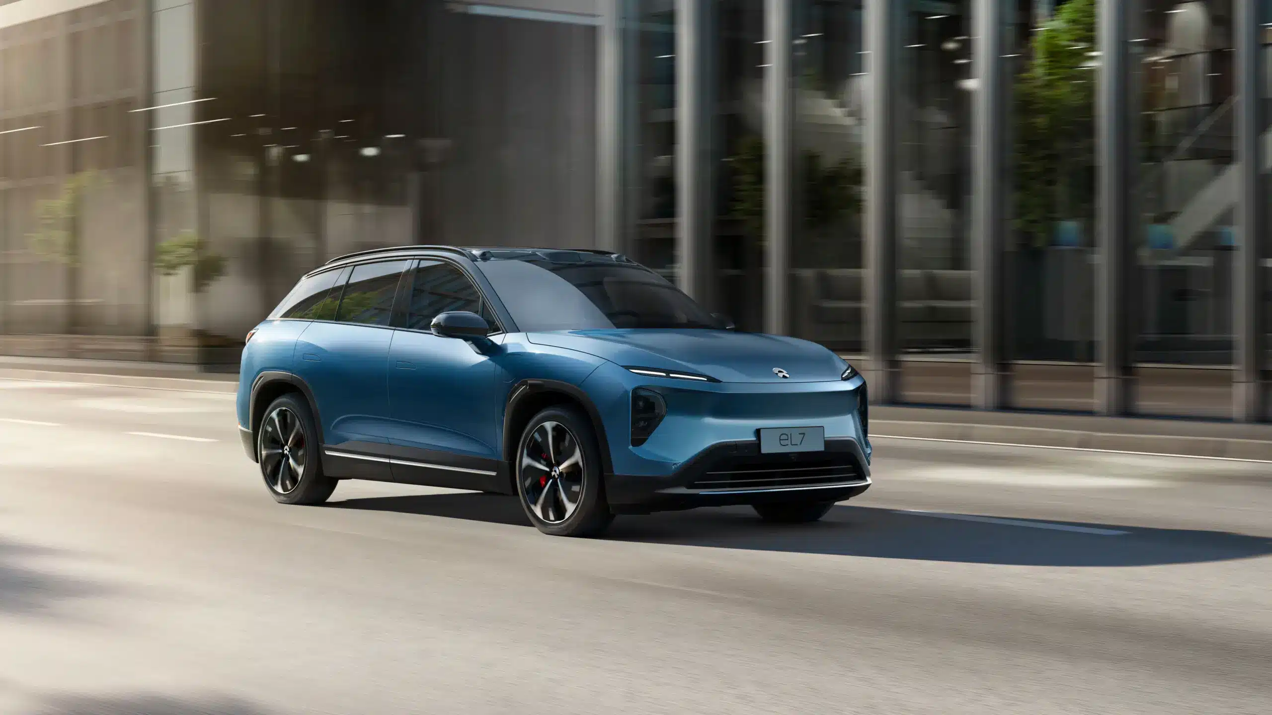 Blauer NIO EL7 SUV fährt auf einer städtischen Straße vor einem Gebäude mit Glasfassade.