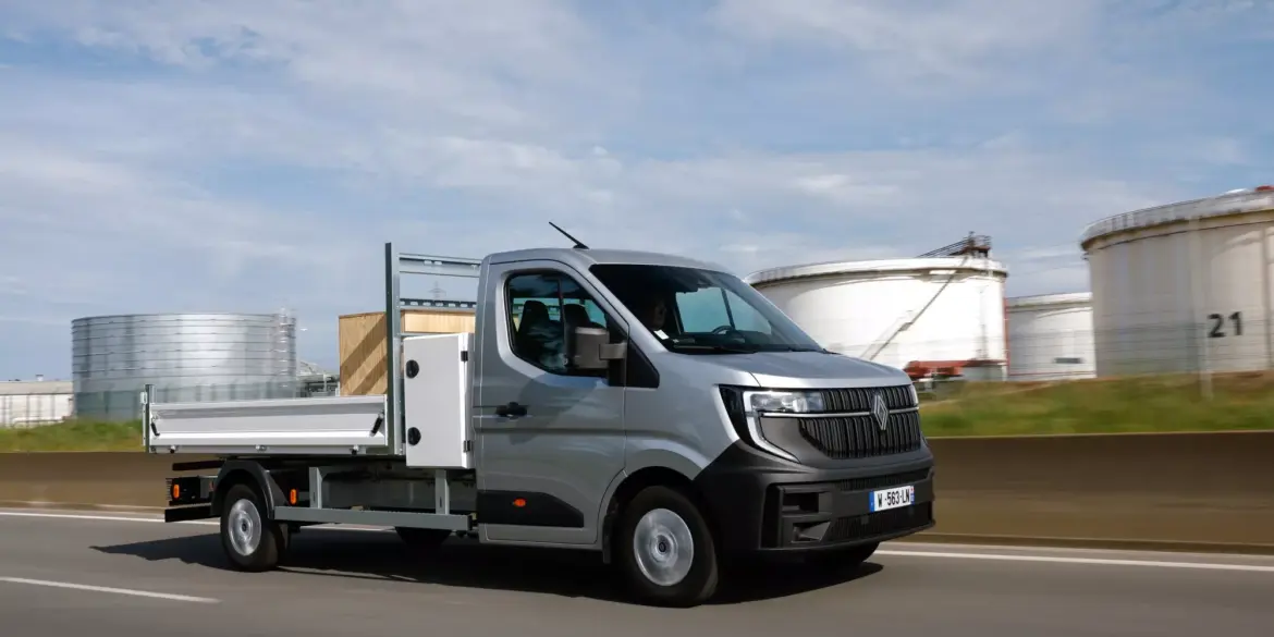 Renault Master Pritschenwagen in Fahrt auf einer Landstraße vor Industrieanlagen.
