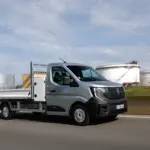 Renault Master Pritschenwagen in Fahrt auf einer Landstraße vor Industrieanlagen.