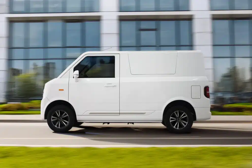 Weißer TX1-e Elektro-Transporter fährt auf einer städtischen Straße vor einem modernen Bürogebäude