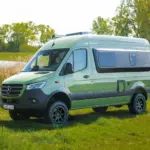 Grüner Westfalia James Cook 600 D Kastenwagen auf Mercedes Sprinter Basis steht auf einer Wiese vor Bäumen und einem See.