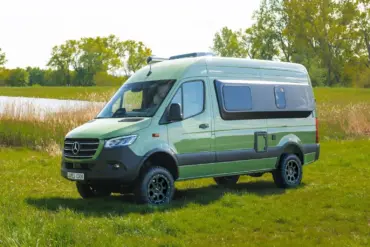 Grüner Westfalia James Cook 600 D Kastenwagen auf Mercedes Sprinter Basis steht auf einer Wiese vor Bäumen und einem See.