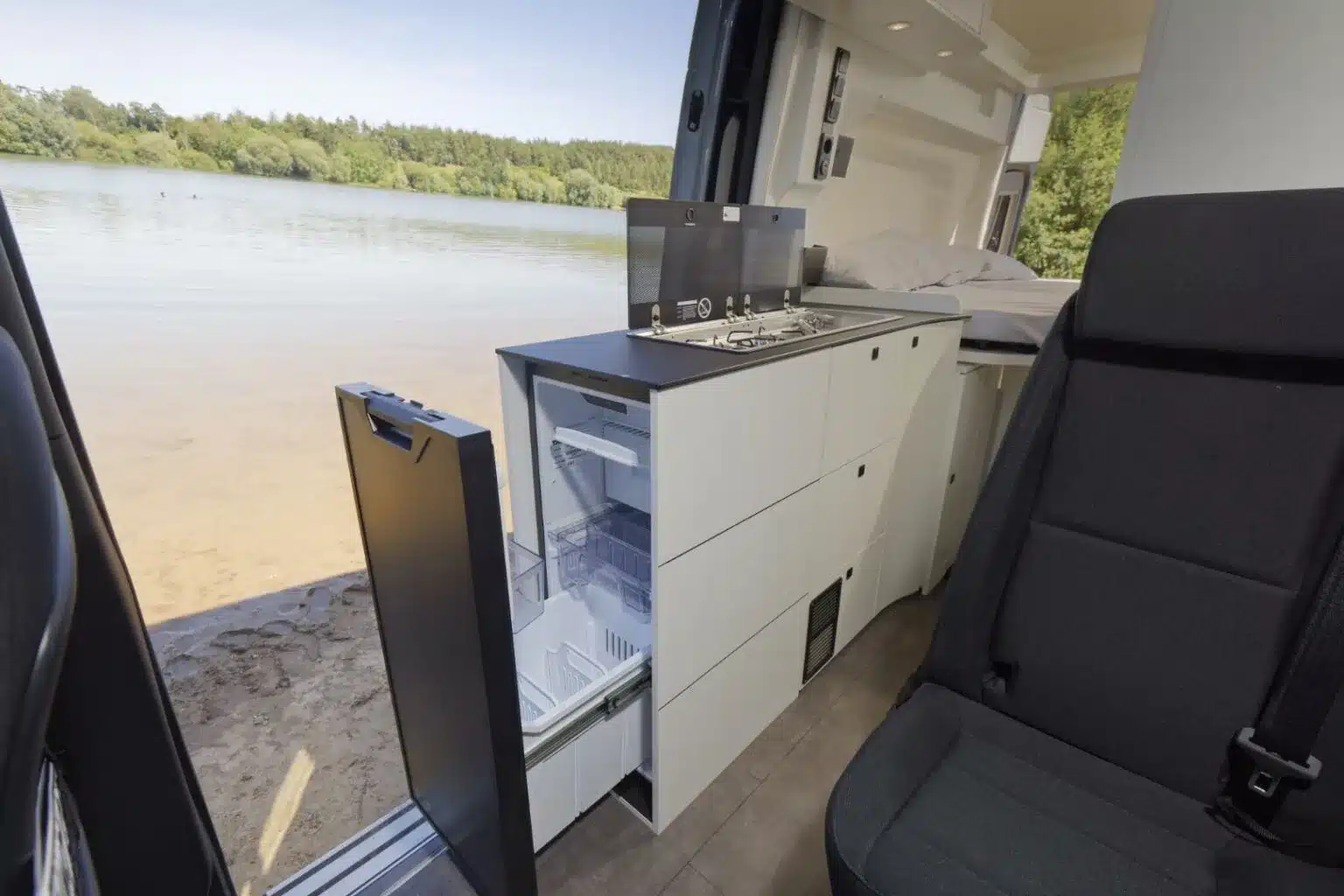 Innenaufnahme der Küche im Westfalia James Cook 600 D mit offenem Kühlschrank und Blick auf das Seeufer.