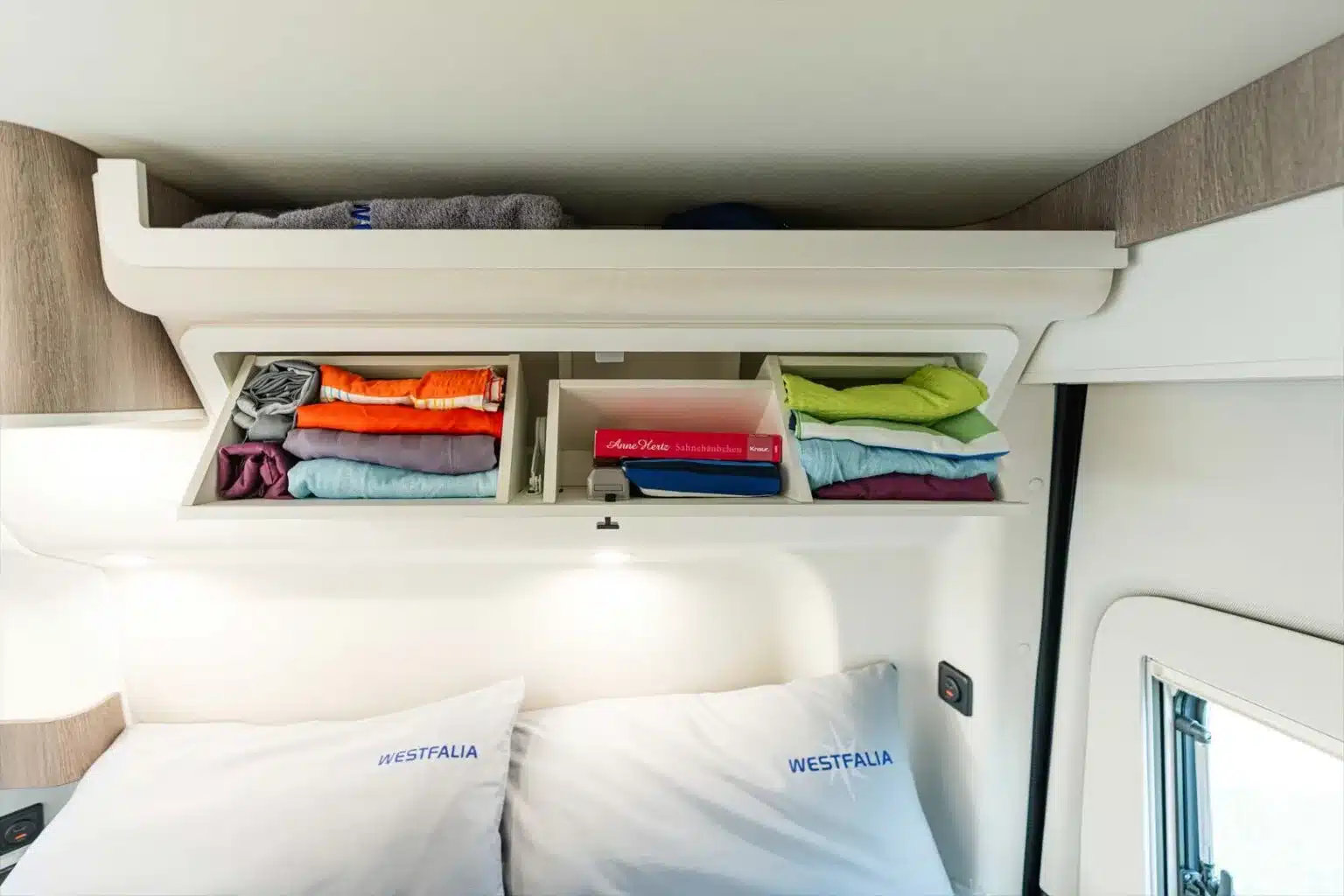 Oberschrank über dem Bett im Westfalia James Cook 600 D, gefüllt mit Kleidung.