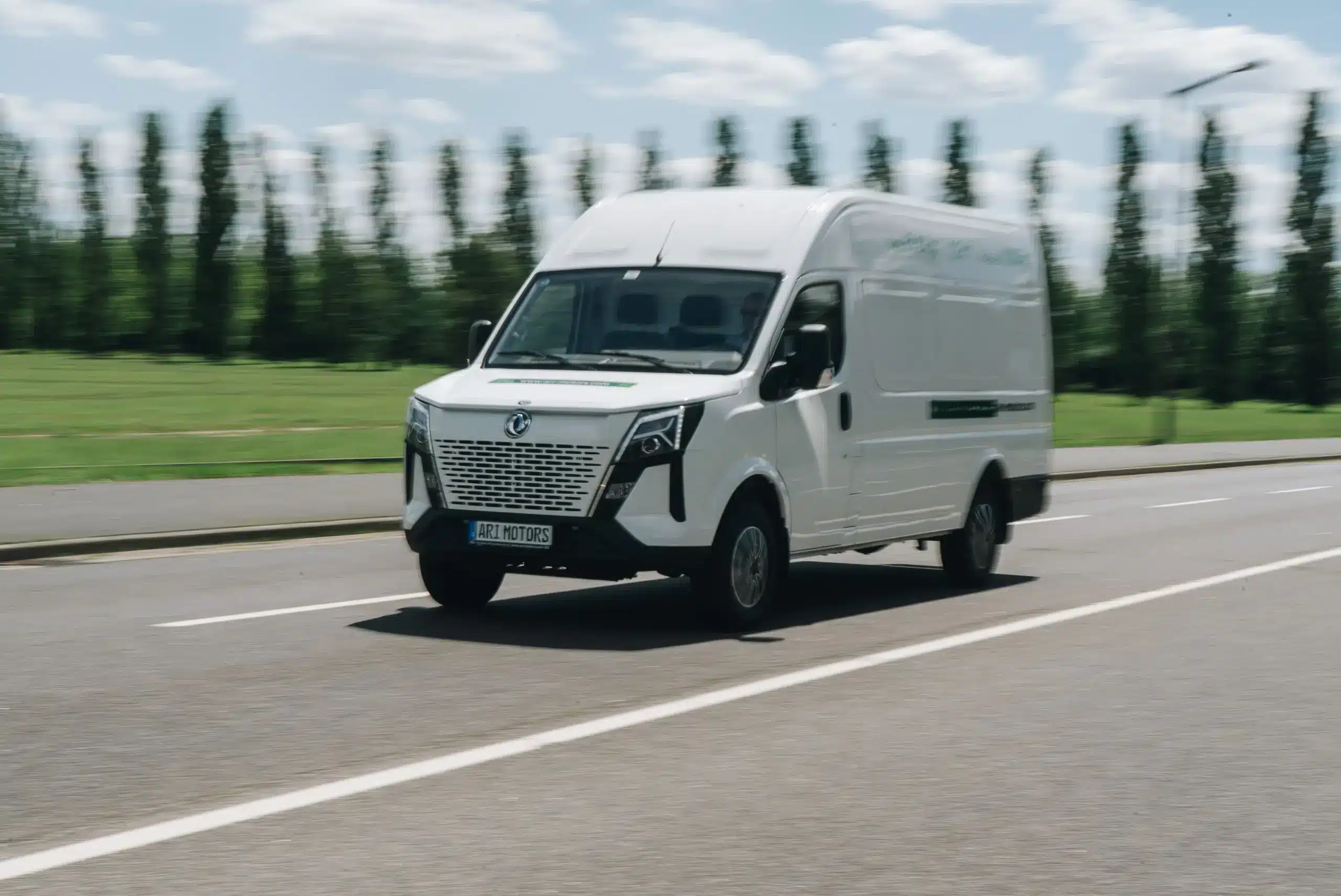 Weißer ARI 1710 Elektro-Transporter fährt auf einer Landstraße, Bäume und Wiese im Hintergrund.