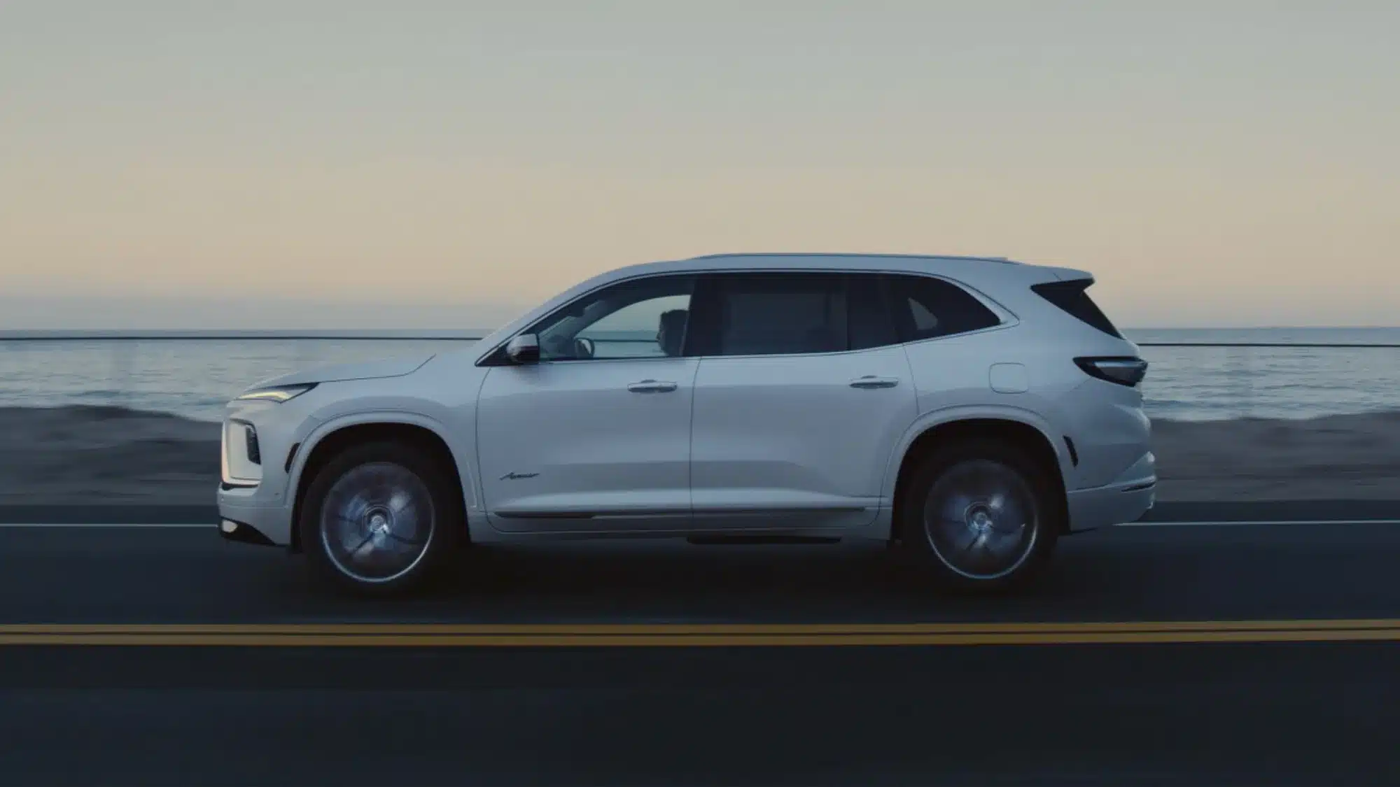 Seitenansicht eines weißen 2026 Buick Enclave auf einer Küstenstraße bei Sonnenuntergang