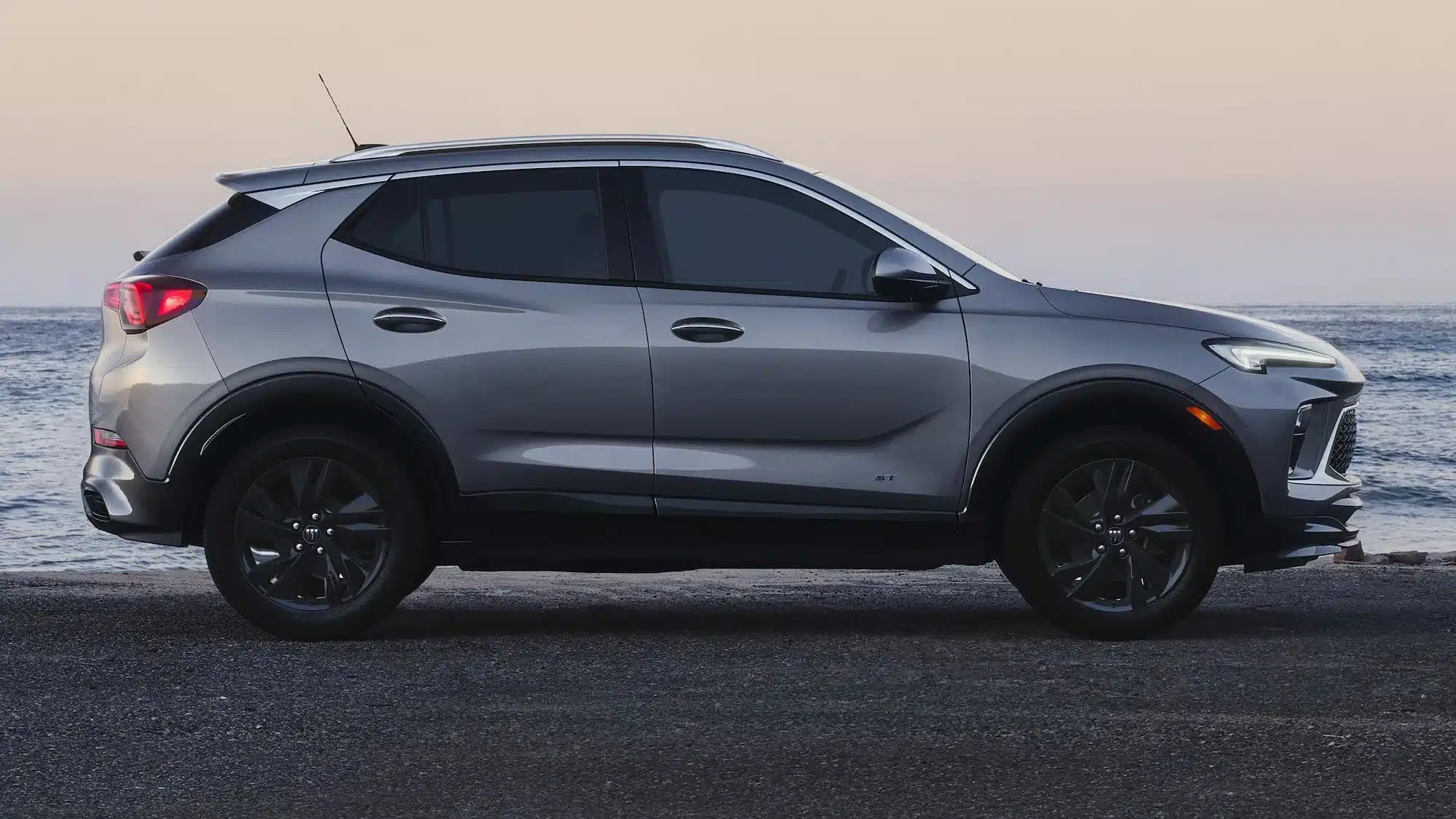 Seitenansicht eines silbergrauen 2026 Buick Encore GX SUV auf einer Küstenstraße vor dem Meer.
