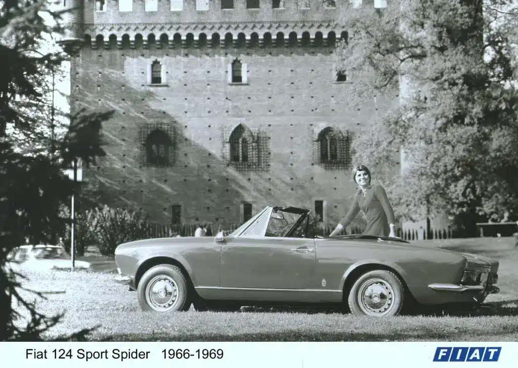 Schwarzweißfoto eines Fiat 124 Sport Spider Cabriolets der ersten Generation (1966-1969), seitlich vor einer Burg, mit einer Frau am Fahrzeug.