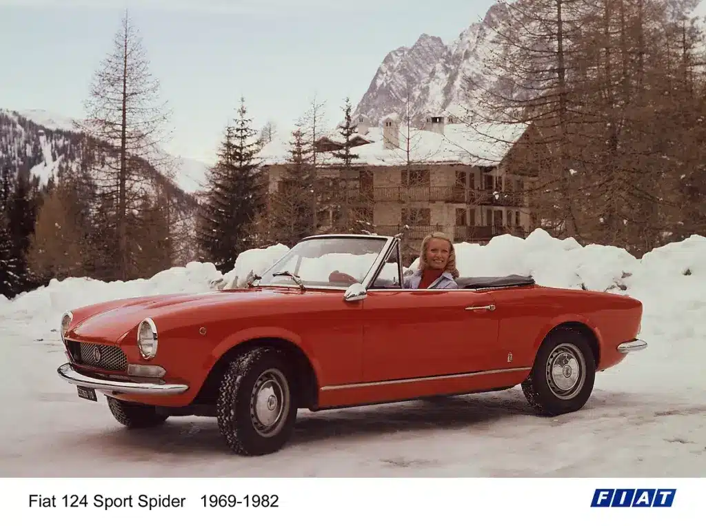 Fiat 124 Sport Spider Cabrio in Rot im Schnee, mit einer Frau am Steuer, vor alpiner Winterkulisse.