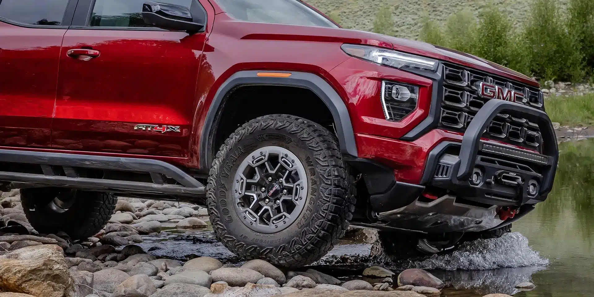 Seitenansicht eines roten GMC Canyon AT4X 2026, der auf felsigem Terrain durch ein seichtes Gewässer fährt.