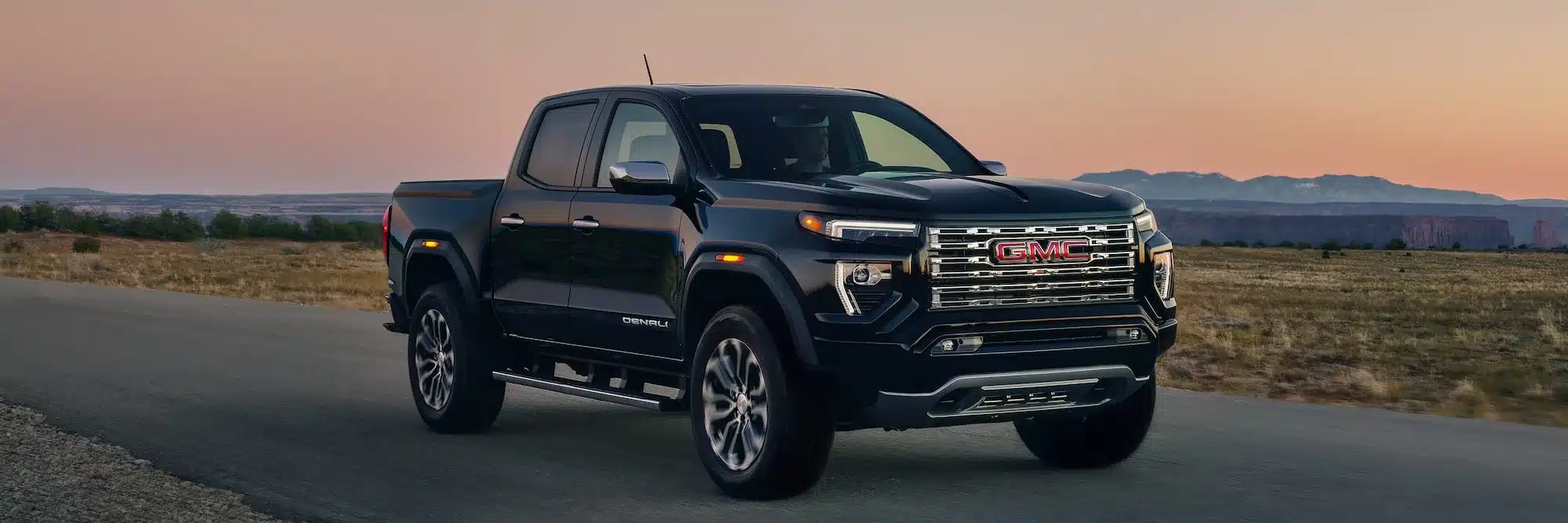 Frontansicht des schwarzen GMC Canyon Denali 2026 im Freien bei Sonnenuntergang, fahrend auf Asphalt mit weiter Landschaft im Hintergrund.