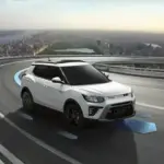 Weißer KGM Tivoli SUV mit schwarzen Felgen fährt auf einer Straße mit Stadtsilhouette im Hintergrund.