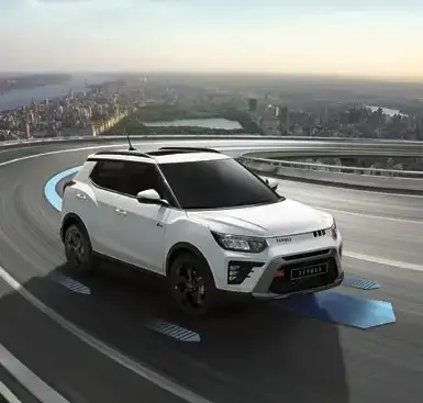 Weißer KGM Tivoli SUV mit schwarzen Felgen fährt auf einer Straße mit Stadtsilhouette im Hintergrund.