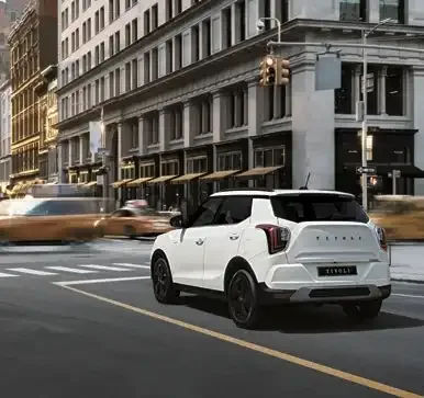 Weißer KGM Tivoli SUV mit schwarzem Dach fährt durch eine Stadt, im Hintergrund sind Gebäude und Taxis zu sehen.