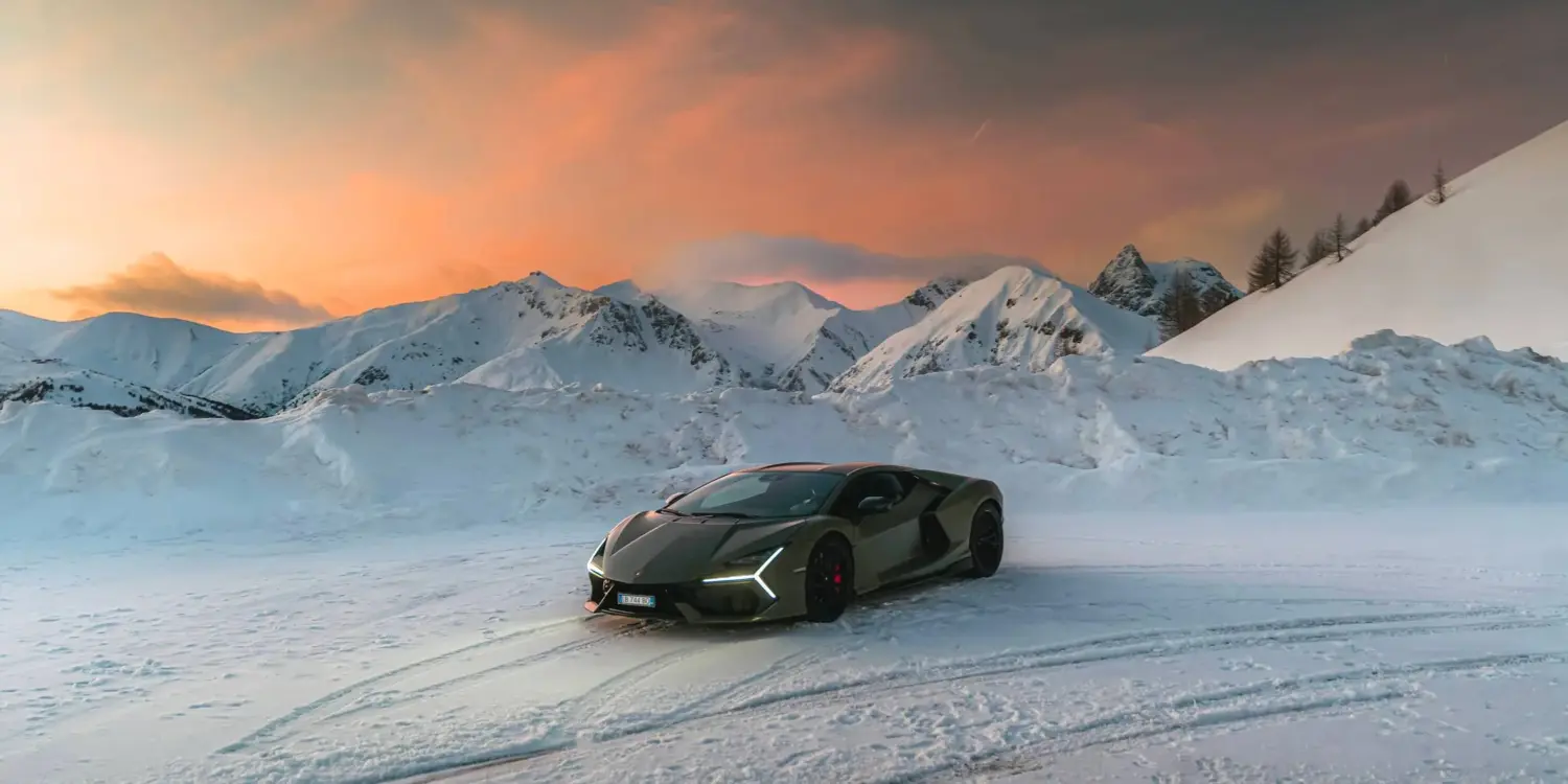 Lamborghini Revuelto steht auf Schnee vor schneebedeckten Bergen bei Sonnenuntergang.