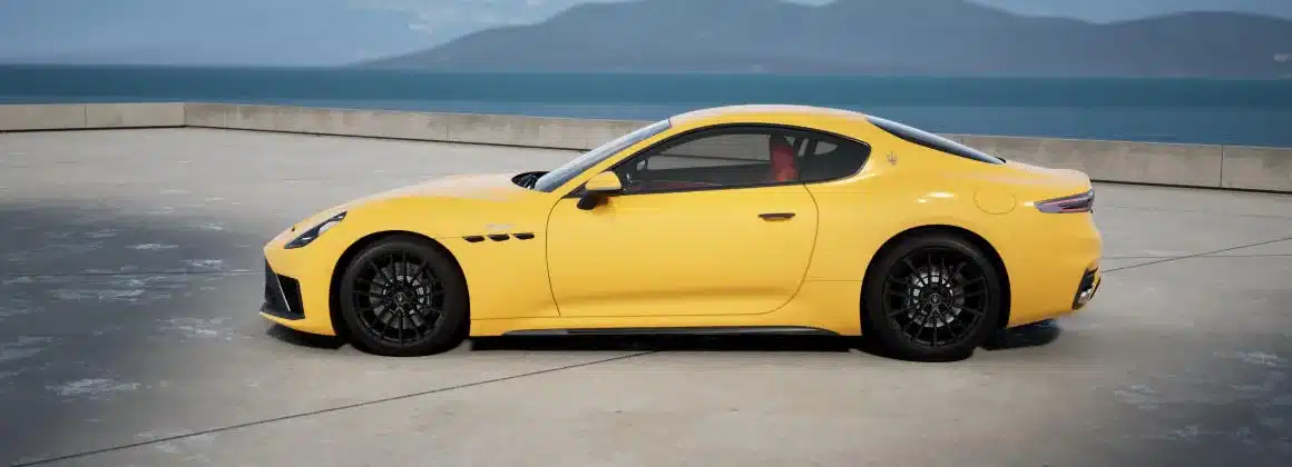 Gelber Maserati GranTurismo Trofeo 2027 als Coupé, Seitenansicht auf Betonfläche mit Wasser und Bergen im Hintergrund.