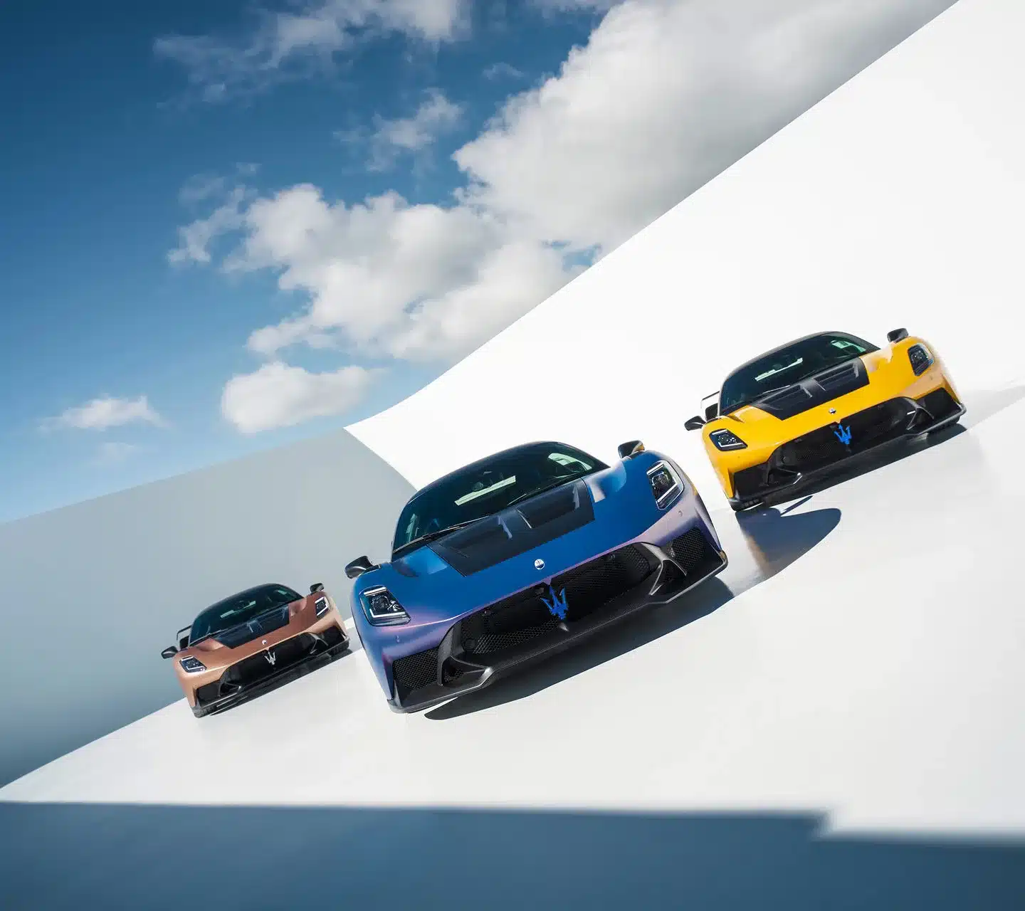 Drei Maserati GT2 Stradale Sportwagen nebeneinander, vorne in Blau, rechts in Gelb und links in Kupferbraun, auf weißem Untergrund mit blauem Himmel