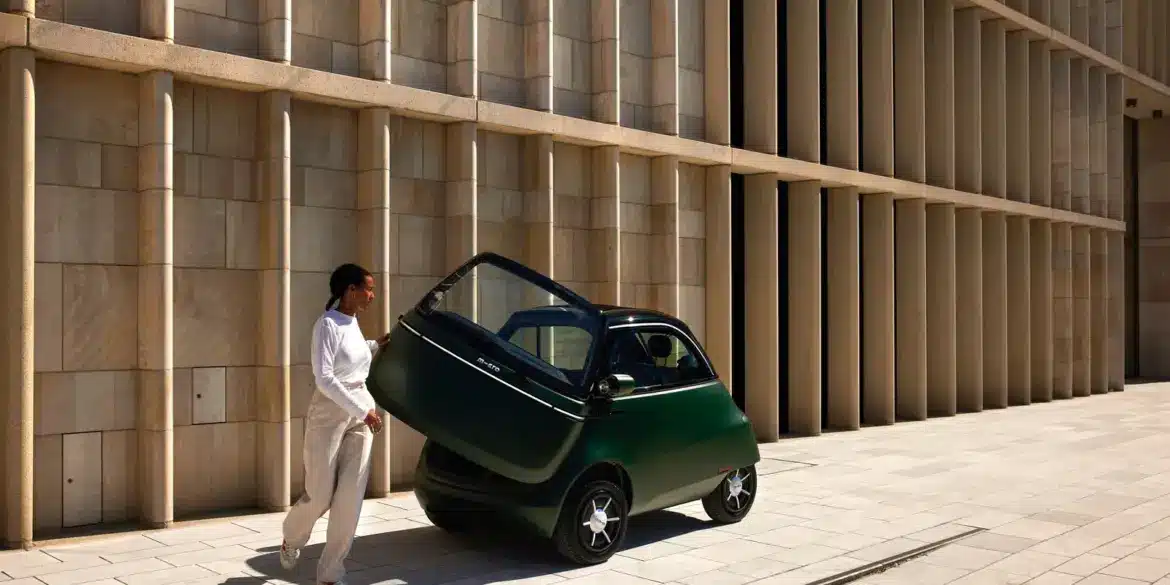 Grüner Microlino mit geöffneter Fronttür, eine Person steigt aus, moderne Gebäudewand im Hintergrund.