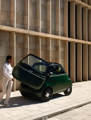 Grüner Microlino mit geöffneter Fronttür, eine Person steigt aus, moderne Gebäudewand im Hintergrund.