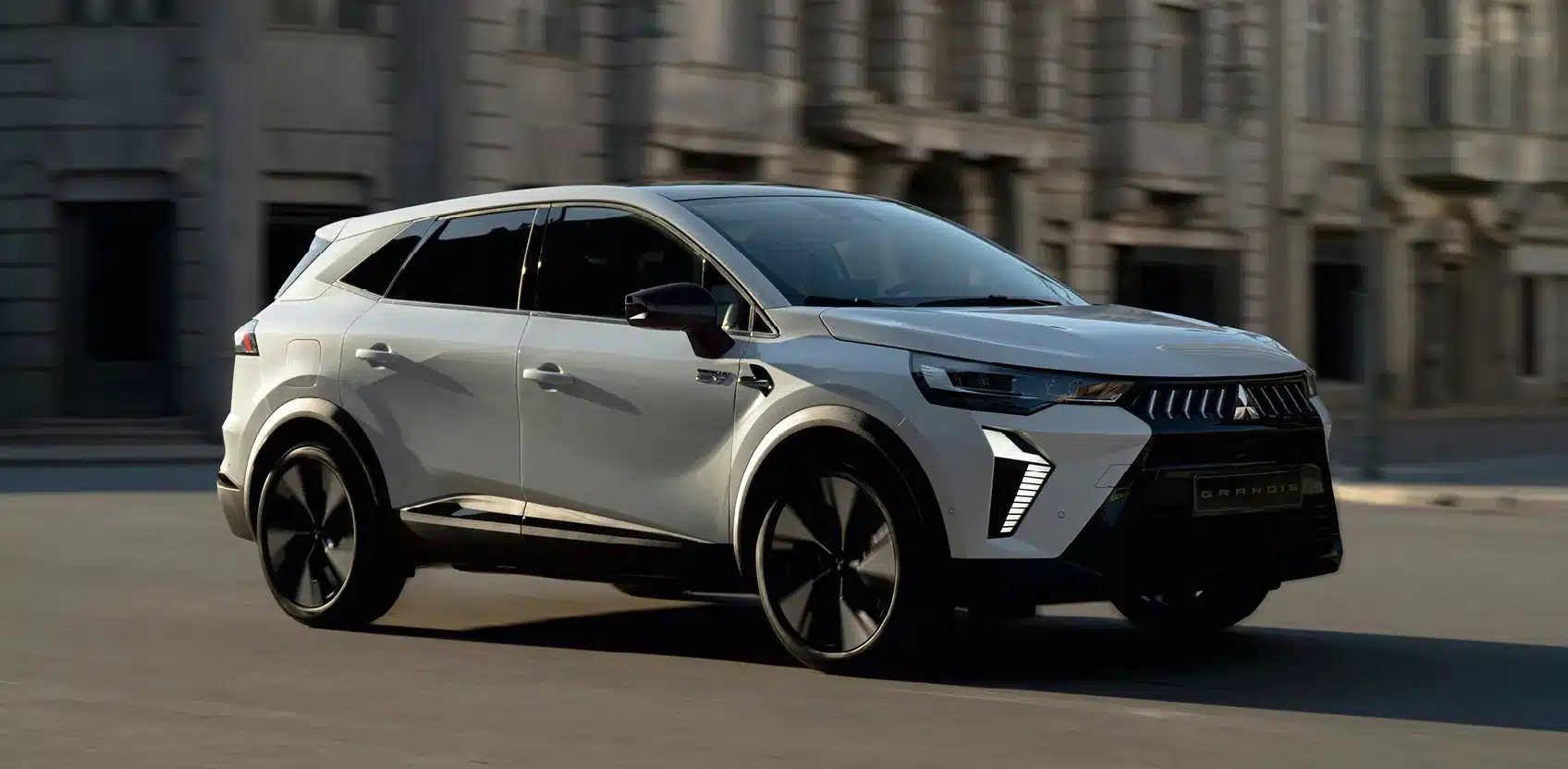 Mitsubishi Grandis 2024 als weißer Hybrid-SUV fährt vor urbaner Kulisse.