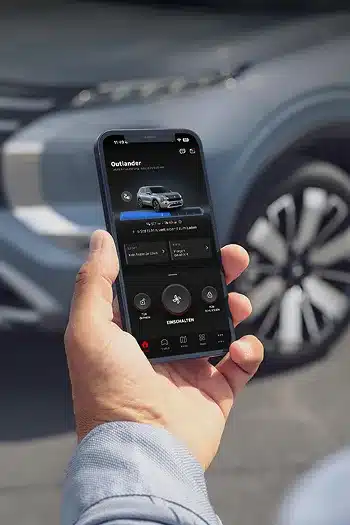 Hand hält Smartphone mit geöffneter Mitsubishi Motors App vor dem Outlander PHEV.