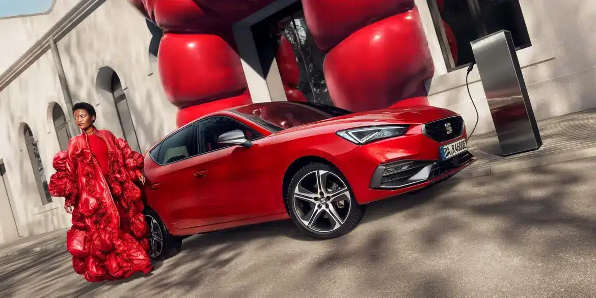 Roter SEAT Leon e-HYBRID beim Laden an einer Ladestation vor einem modernen Gebäude, daneben eine Frau in auffälligem roten Outfit.