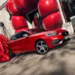 Roter SEAT Leon e-HYBRID beim Laden an einer Ladestation vor einem modernen Gebäude, daneben eine Frau in auffälligem roten Outfit.