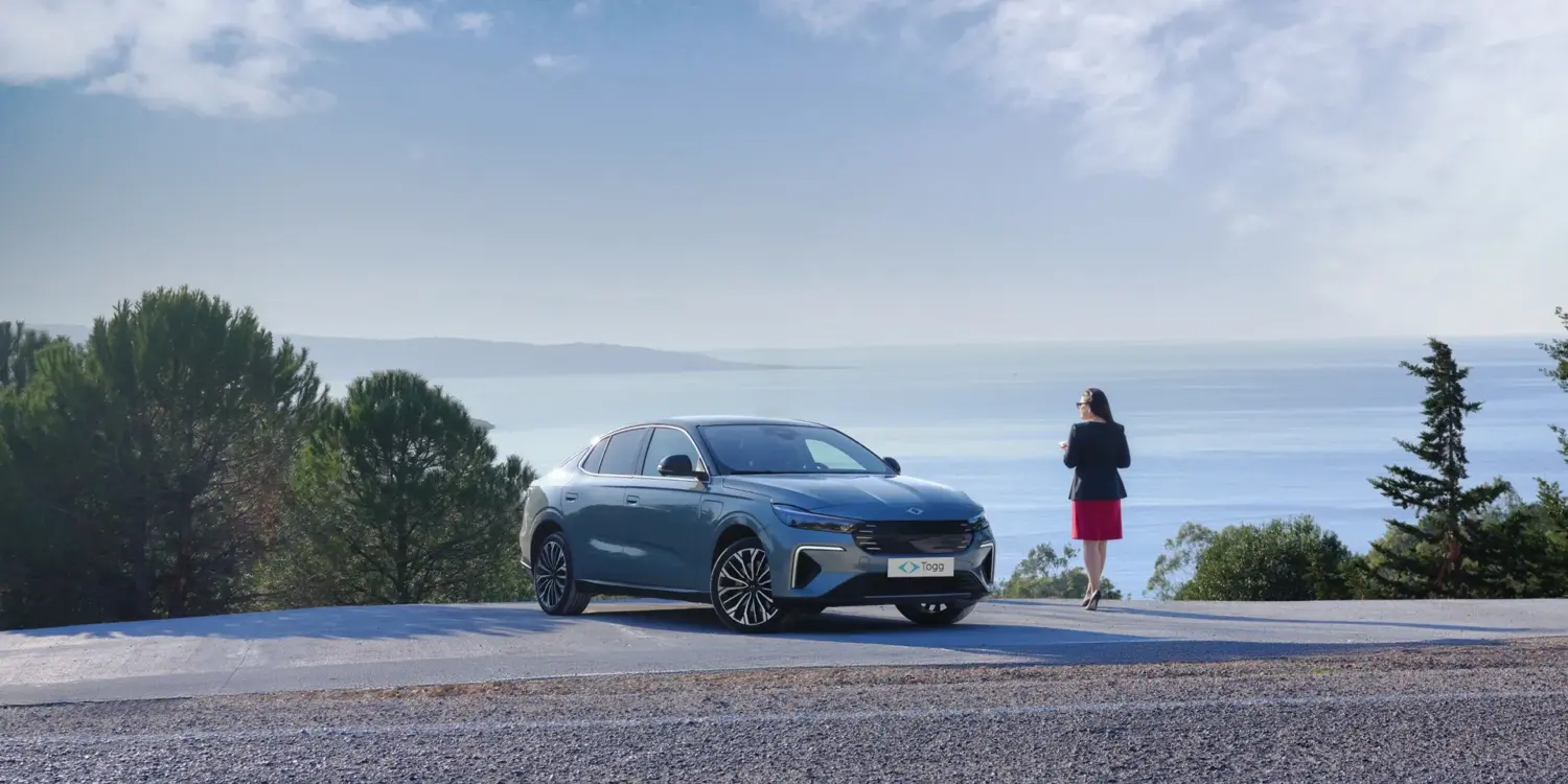 Der neue Togg T10F Fastback steht auf einer Straße mit Blick aufs Meer, daneben eine Frau, Bäume im Hintergrund.