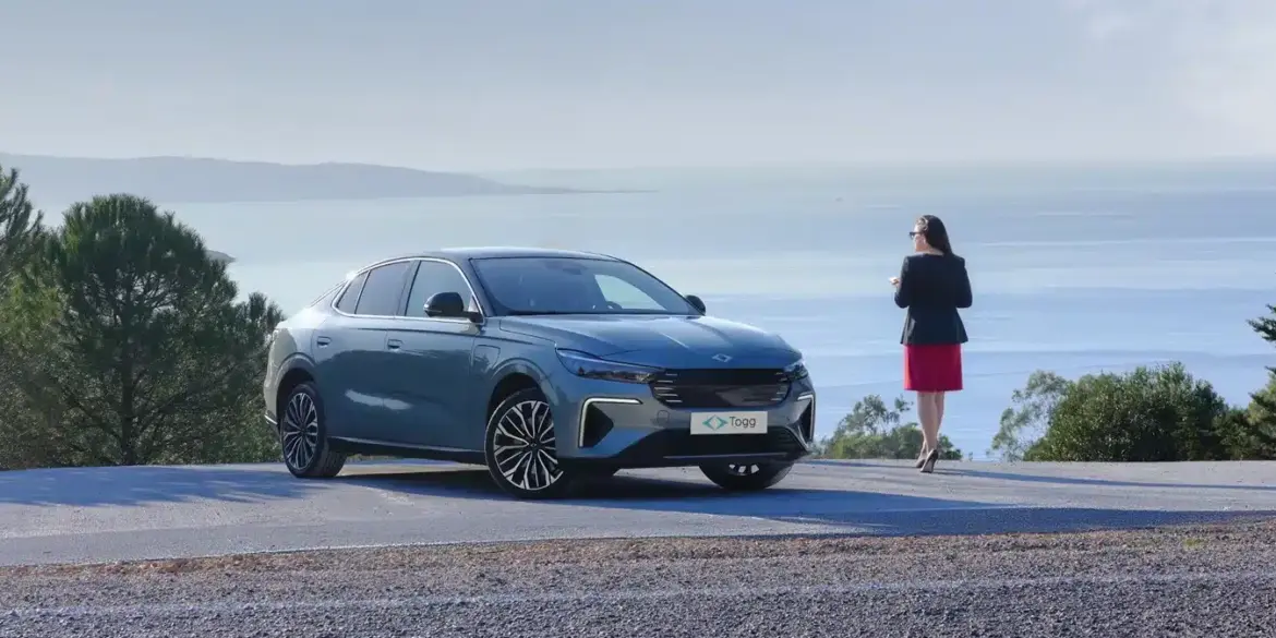Der neue Togg T10F Fastback steht auf einer Straße mit Blick aufs Meer, daneben eine Frau, Bäume im Hintergrund.